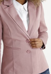 Blazer rosa chiaro realizzato in tessuto testurizzato, con colletto a riscottura, due bottoni e una tasca laterale. Indossato sopra una camicia bianca.