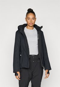 Čierna bunda s kapucňou, vybavená predným zipsom, veľkými vreckami a dlhými rukávmi. Nosená cez sivé tričko a čierne nohavice. Hladká textúra.