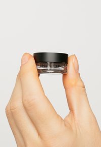 Petit pot transparent avec un couvercle noir, contenant une substance opaque. Le pot a une texture lisse et un minimum de branding sur le côté.