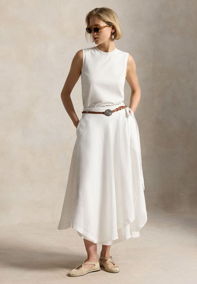 Moteris vilki baltą rankovių neturinčią suknelę su rudu diržu, dėvi balkšvus batelius ir akinius nuo saulės, stovi prieš neutralią smėlio spalvos foną.