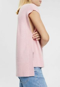 Pull sans manches rose avec une texture côtelée, une coupe décontractée et des fentes latérales, associé à un denim bleu clair, présentant un design décontracté.