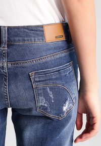 Jeans de mezclilla en azul medio con un acabado desgastado, que presentan una etiqueta de cuero marrón en la parte trasera de la cinturilla y un diseño de bolsillo trasero curvado.