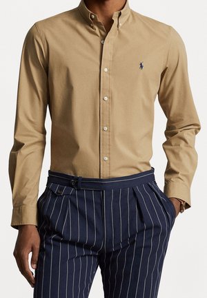 Homme portant une chemise beige boutonnée à manches longues avec un petit logo et un pantalon bleu marine à fines rayures avec détail ceinture et mains dans les poches.