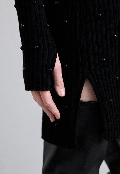 Maglione nero a coste con perle nere rotonde, caratterizzato da uno spacco laterale e maniche lunghe, abbinato a pantaloni in pelle scuri.