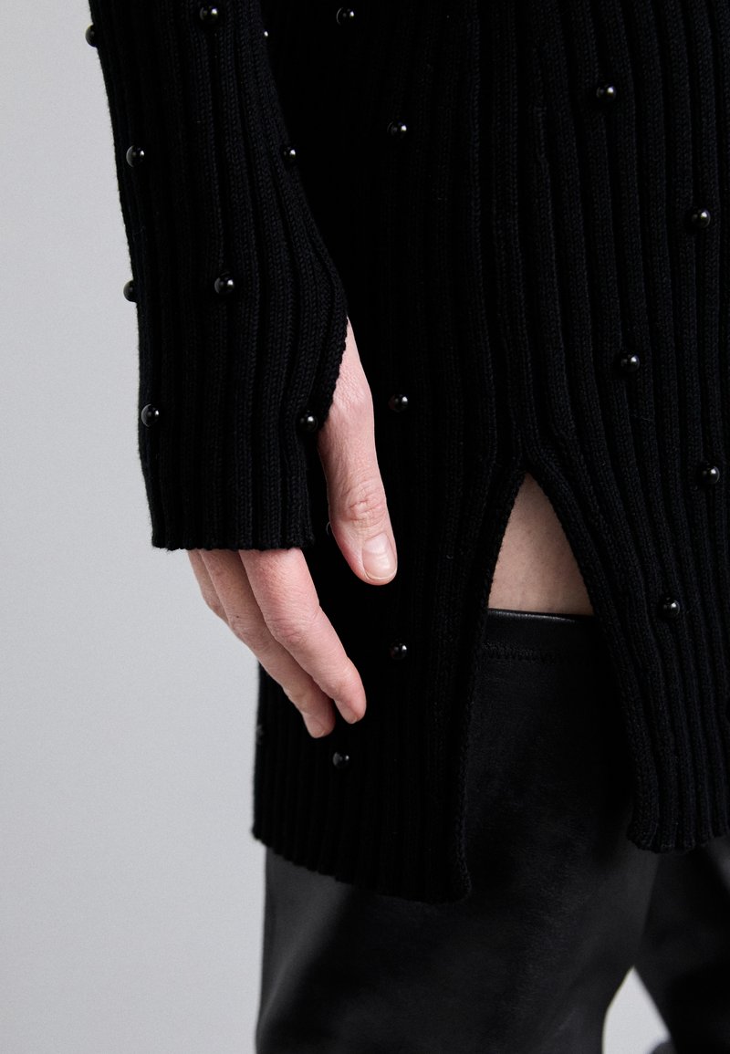 Pull en maille côtelée noire avec des perles noires rondes, présentant une fente sur le côté et des manches allongées, associé à un pantalon en cuir sombre.