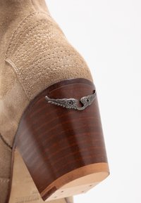 Beżowe zamszowe botki za kostkę z drewnianym obcasem, z metalicznym akcentem w kształcie skrzydła z tyłu. Teksturowana cholewka i gładkie wykończenie obcasa.