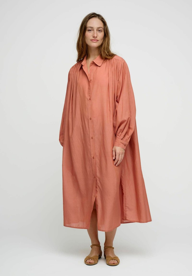 Femme debout portant une robe midi corail ample à manches longues boutonnée avec des sandales beige ouvertes à l’avant sur un fond blanc uni.