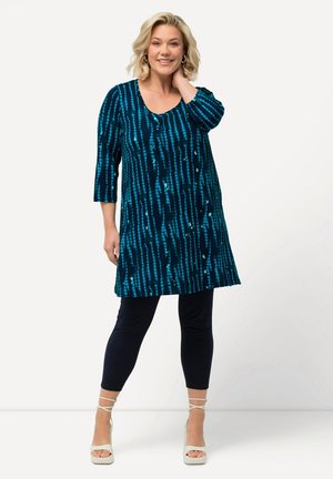 Tunique teal avec des rayures verticales bleues et des manches trois-quarts, associée à des leggings navy et des talons blancs à lanières.