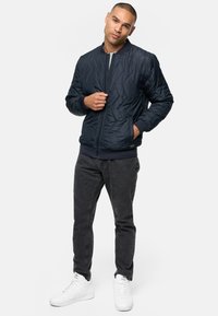 Chaqueta bomber de algodón de marina con un diseño acolchado, puños y dobladillo de canalé, cierre frontal con cremallera, dos bolsillos laterales, combinada con pantalones vaqueros negros y zapatillas blancas.