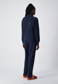 Tuta blu navy con colletto alto, polsini a coste e pantaloni dritti. Indossato con scarpe rosse con un logo bianco. Tessuto liscio.