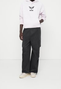 Person klädd i en ljusrosa huvtröja med en fjäril och japansk text, mörka vida cargobyxor och krämfärgade sneakers som står framför en vit bakgrund.