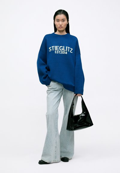 Plavi oversized pleteni pulover s natpisom "STIEGLITZ EST2014" u krem boji, uparen s svijetloplavim trapericama s izrezom i sjajnom crnom torbom.