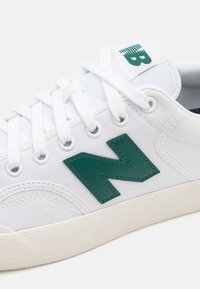 Zapatilla blanca con un upper de lona texturizada, logo "N" de cuero verde, cordones blancos y suela de goma. Diseño sencillo con acentos mínimos.