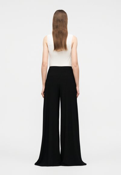 Γυναίκα με μακριά καστανά μαλλιά που φοράει λευκή αμάνικη μπλούζα και μαύρο παντελόνι με φαρδιά μπατζάκια, στέκεται με την πλάτη γυρισμένη σε απλό φόντο.