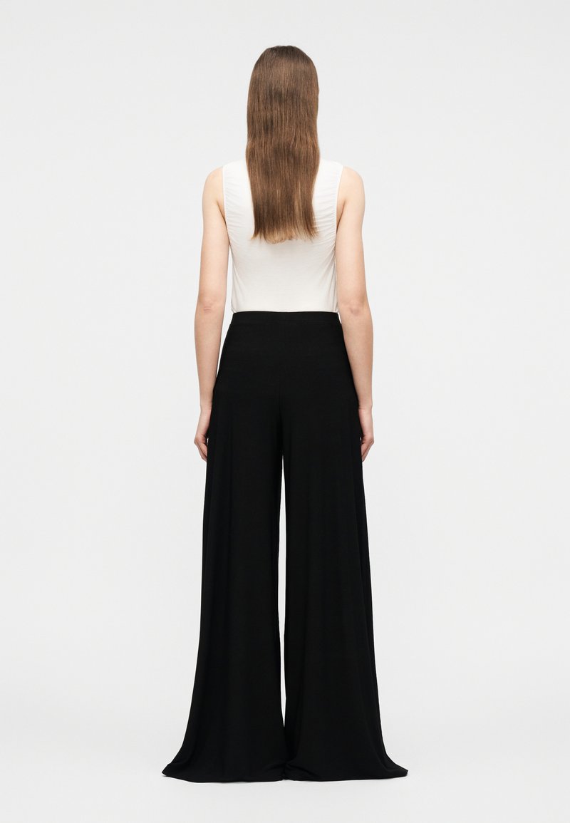 Femme aux longs cheveux bruns, portant un haut blanc sans manches et un pantalon noir à jambes larges, debout de dos sur un fond uni.