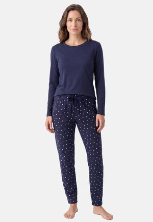 Femme debout pieds nus, portant un haut à manches longues bleu marine et un pantalon de pyjama bleu marine avec de petits motifs de cœurs blancs.