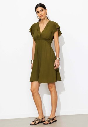 Femme portant une robe vert olive à col en V avec des manches volantées, des bracelets dorés, des boucles d’oreilles créoles, et des sandales à imprimé léopard, debout contre un mur blanc.