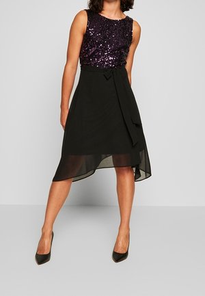 Mujer con vestido negro sin mangas con parte superior de lentejuelas moradas y bajo asimétrico transparente, combinado con tacones altos negros.