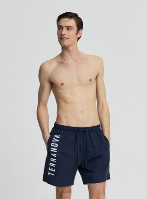 Giovane uomo in piedi senza maglietta con le mani in tasca, indossa pantaloncini da bagno blu navy con la scritta "TERRANOVA" stampata verticalmente sulla gamba sinistra.