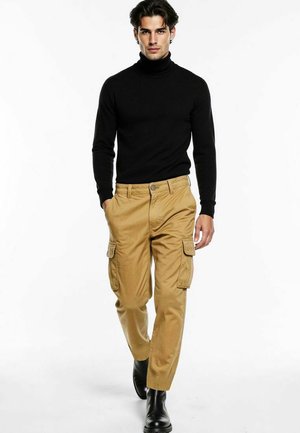 Hombre caminando vestido con suéter negro de cuello alto, pantalones cargo color canela y botas negras sobre fondo blanco.