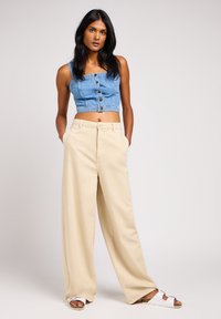 Top corto de denim con detalles de botones, combinado con pantalones beiges de corte amplio. Modelo usando sandalias blancas de plataforma. Fondo liso.