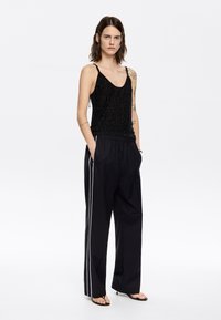 Schwarzes Pailletten-Trägershirt mit dünnen Trägern, kombiniert mit lockeren schwarzen Hosen mit weißen Seitenstreifen. Glatte Textur, entspannter Schnitt.