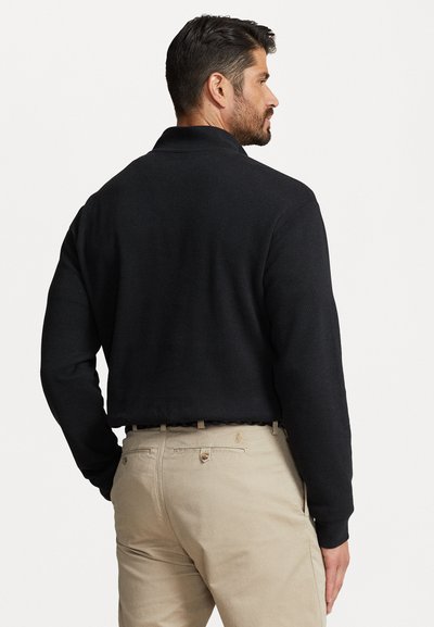 Black sweater with a collar and long sleeves, made of textured fabric, paired with khaki pants featuring a logo on the back pocket.