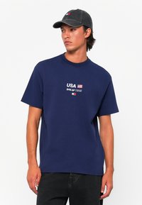 Navy blue cotton t-shirt featuring “USA” and flag graphics. Short sleeves, crew neck, relaxed fit. Paired with a dark denim cap.