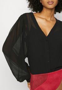 Blouse noire avec manches longues transparentes et bouffantes, encolure en V et boutons sur le devant. Présente une texture lisse, associée à une jupe en daim rouge.