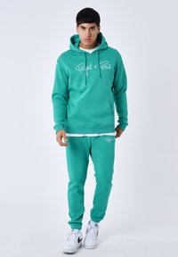 Sudadera color turquesa y pantalones de chándal a juego, hechos de un material suave, con un bolsillo frontal y logotipo en blanco. Estilizados con zapatillas blancas.