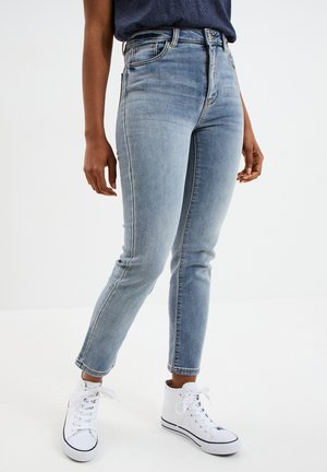 Person, die hellblaue Slim-Fit-Jeans und weiße High-Top-Sneakers trägt, steht vor einem schlichten weißen Hintergrund.