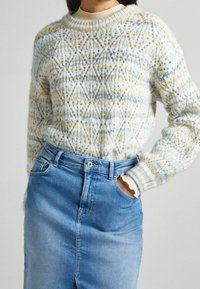 Maglione lavorato a maglia con motivo geometrico in blu chiaro, crema e beige; scollo rotondo e polsini a costine abbinati a una gonna di jeans.