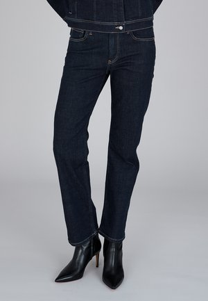 Osoba nosząca ciemnoniebieskie, proste dżinsy oraz czarne botki na wysokim obcasie z ostrym noskiem na jednolitym tle.