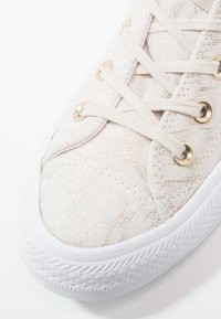 Jasno beżowe sneakersy o teksturowanej powierzchni, z delikatnym metalicznym połyskiem i białą gumową podeszwą. Posiadają złote oczka i kremowe sznurowadła.