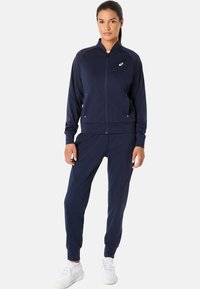 Marine blauwe trainingspak van glad, rekbaar materiaal; bevat een een ritsjack, aflopende broek en zijzakken; logo op de borst.