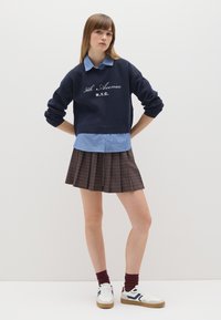 Marineblauer Pullover mit gestickter Aufschrift "5th Avenue N.Y.C.", darüber ein hellblau gestreiftes Hemd. Kombiniert mit einem braunen karierten Faltenrock und weißen Sneakers.