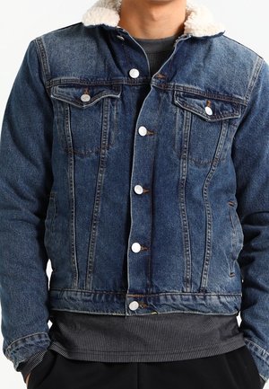 Homme portant une veste en jean bleue avec un col sherpa blanc et des boutons argentés sur un pull côtelé foncé.