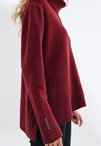 Bordowy sweter z wysokim kołnierzem; miękka tekstura, prążkowane akcenty na rękawach i dołach, boczne rozcięcia oraz metka z logo na mankiecie.