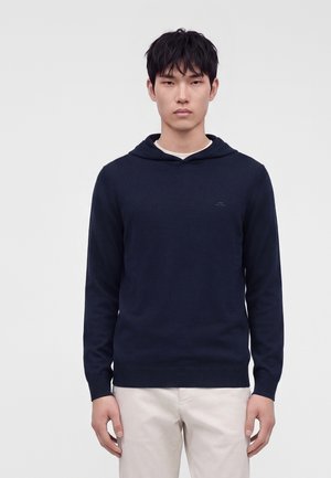 Jeune homme portant un sweat à capuche bleu marine foncé et un pantalon beige clair, debout devant un fond clair uni.