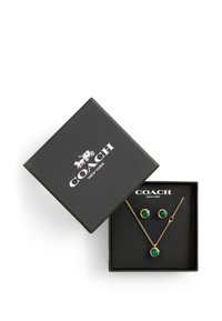 Smyckeset i guldton med örhängen som har runda gröna stenar och ett matchande halsband, presenterat i en svart texturerad box med logotyp.