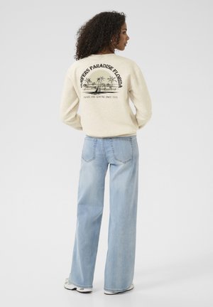 Krämfärgad sweatshirt med tryck "Surfers Paradise Florida" på ryggen. Kombinerad med ljusblå vida jeans och grå sneakers.