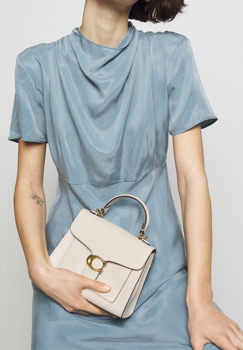 Personne portant une robe bleu clair tenant un petit sac à main beige avec un fermoir doré et une poignée courte, montrant un petit tatouage sur l'avant-bras gauche.