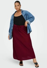 Ženska v črni majici brez rokavov, prevelikem jeans jakni, burgundski plisirani maksi krili, črnih sandalih in zlatih uhani v obliki obroča, samozavestno pozira.