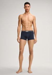 Uomo muscoloso in piedi a piedi nudi, indossando boxer blu navy con elastico griffato, su uno sfondo chiaro e semplice, rivolto frontalmente.