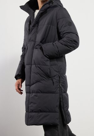 Persona con un abrigo largo negro acolchado con capucha, detalle de cremallera lateral y manos en los bolsillos del abrigo, sobre un fondo liso.