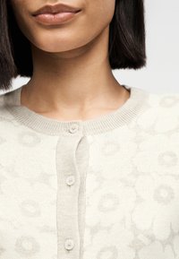 Γυναίκα που φοράει ένα κρεμ πλεκτό ζακέτο με κουμπιά και διακριτικά λουλουδάτα μοτίβα, με ίσια σκούρα μαλλιά και φυσικό χρώμα χειλιών, εμφανιζόμενη από το πηγούνι μέχρι το επάνω μέρος του στήθους.