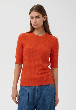 Vrouw met steil bruin haar draagt een oranje trui met korte mouwen en textuur en een blauwe spijkerbroek, staand tegen een effen lichte achtergrond.