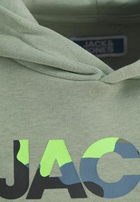 Svetlozelená mikina s kapucňou z textúrovaného materiálu, na ktorej sa nachádzajú výrazné čierne a maskáčové písmená "JAC" s neónovými žltými a modrými akcentmi.