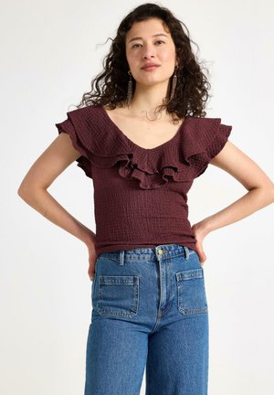 Bordeaux-färgad smockad blus med volangärmar, tillsammans med högmidjade blå denimjeans. Strukturen är krinklad och välarbetad.