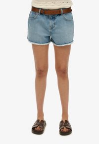Pantalones cortos de mezclilla, de color azul claro con el dobladillo deshilachado, complementados con un cinturón marrón. Sandalias de tiras marrones con un diseño trenzado y suela oscura.